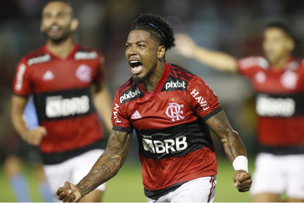 Gabigol viaja com o Flamengo. Jogador aparece em primeiro plano, com uma camisa vermelha com detalhes em preto e branco. Ele está gritando ao comemorar um gol.