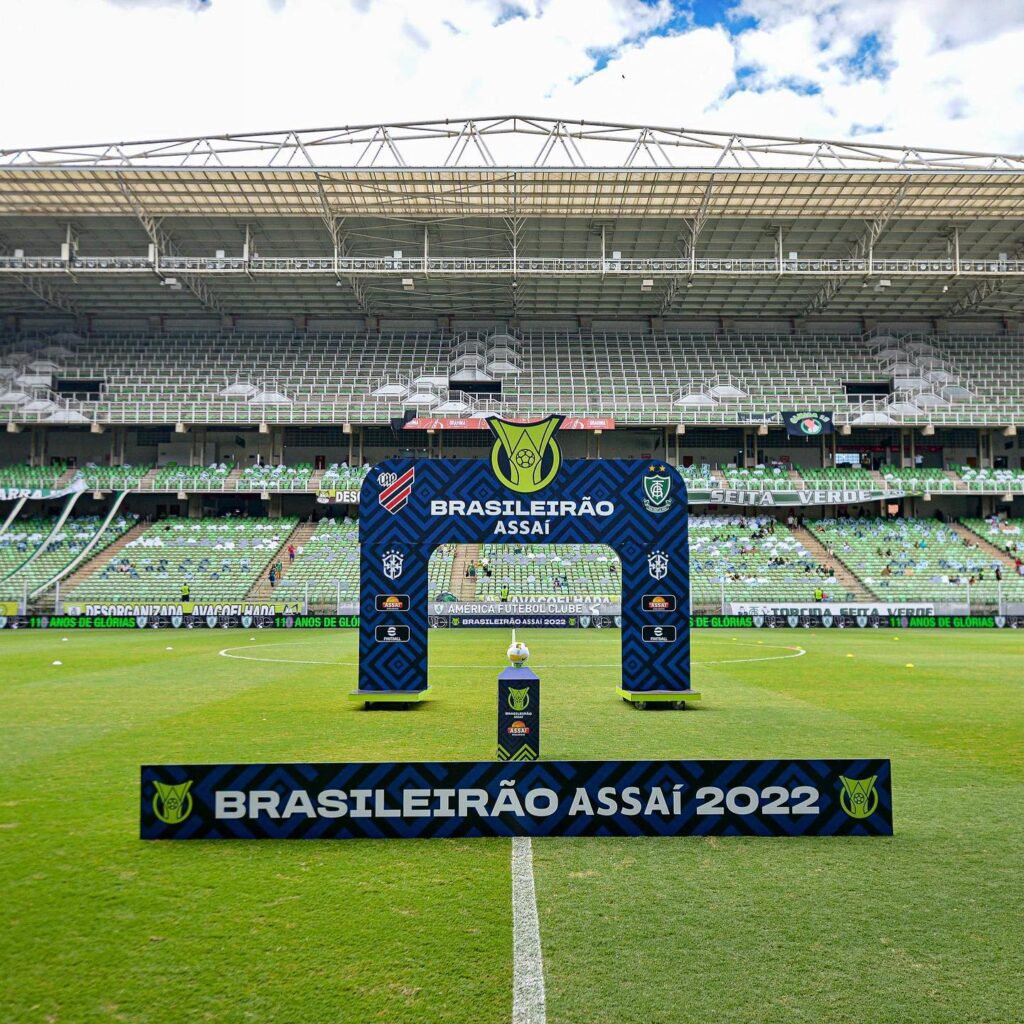 Placa com a frase "Brasileirão Assaí 2022" em frente a totem com a bola do campeonato e, ao fundo, arco da competição, indicando o duelo entre Athletico-PR e América-MG na Arena da Baixada.