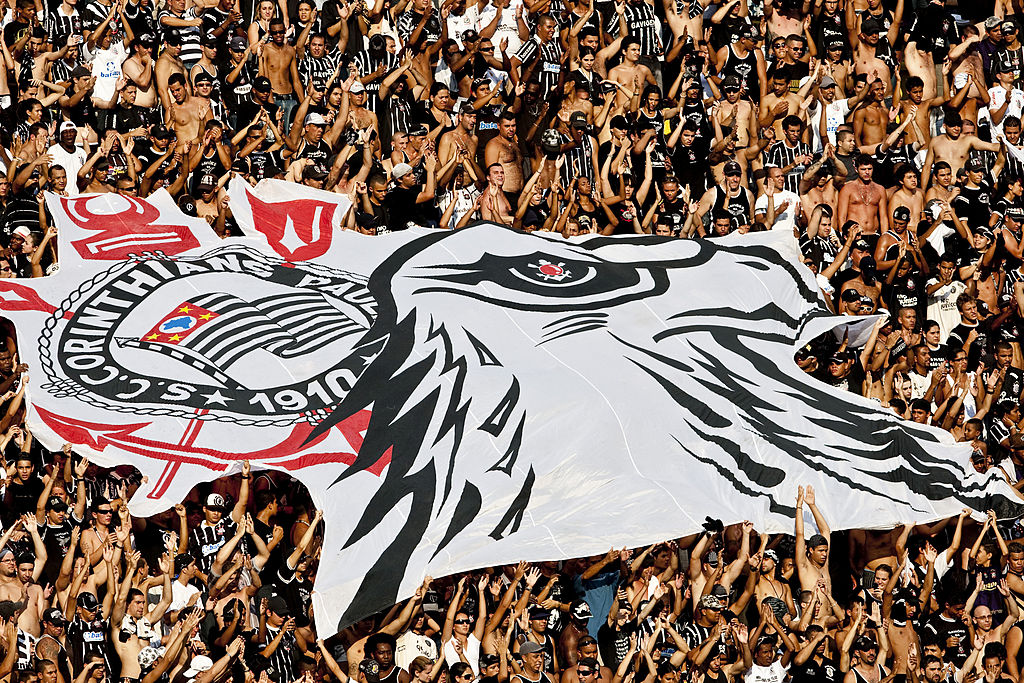 Torcida do Corinthians erguendo bandeirão do escudo do time e de um gavião, mascote do clube.