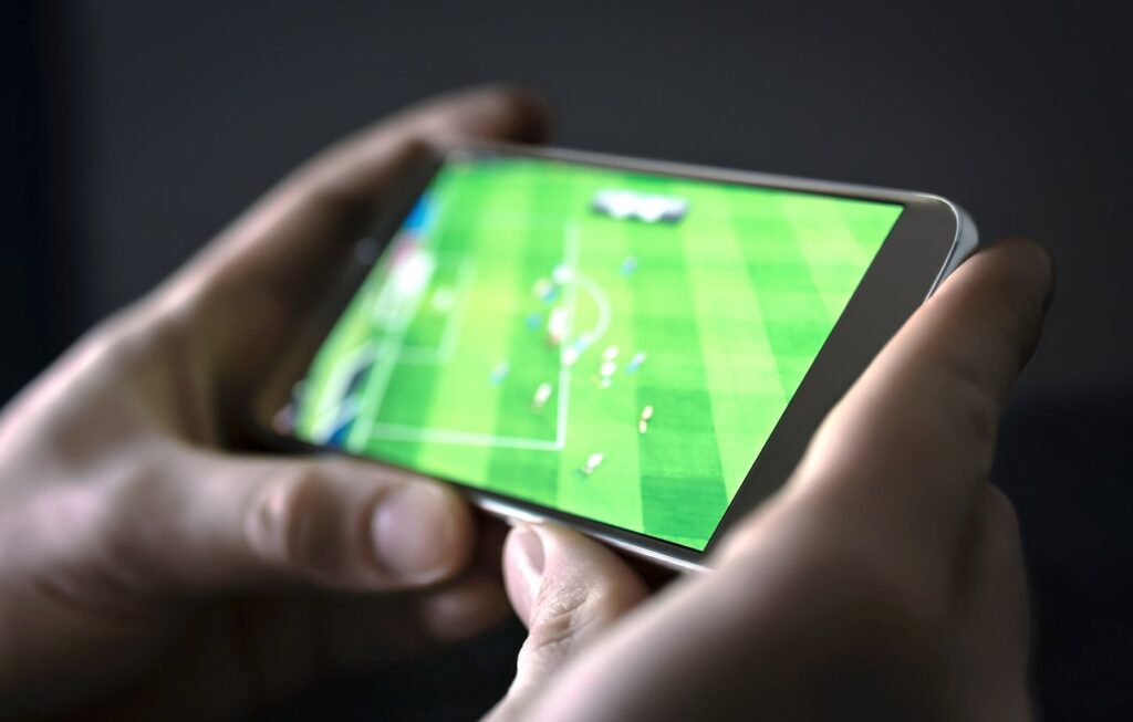 Mãos segurando celular na horizontal e, na tela, partida de futebol sendo transmitida.