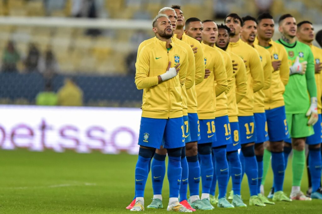Seleção Brasileira perfilada em campo, jogadores com a mão no peito, entoando o hino nacional