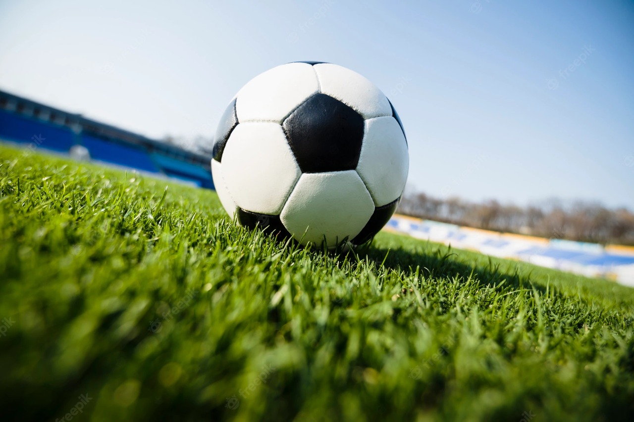 Bola de futebol preta e branca, em cima de gramado de campo de futebol, em imagem levemente inclinada.