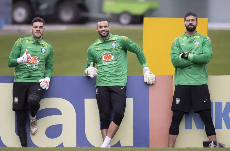 Ederson, Weverton e Alisson, goleiros da Seleção Brasileira, encostados em placa de publicidade na beirada do campo, vestindo uniformes de treino do Brasil