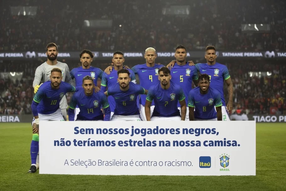 Seleção Brasileira vestindo uniforme azul e posando atrás de placa de campanha contra o racismo no futebol