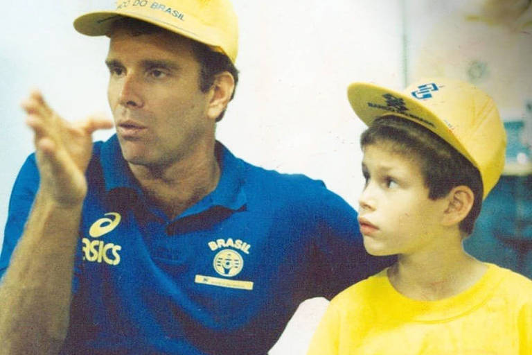 Bernardinho e Bruninho, ainda criança. O pai veste blusa azul da comissão técnica da Seleção Brasileira e vôlei e boné amarelo e o filho veste blusa amarela e boné igual ao do pai.