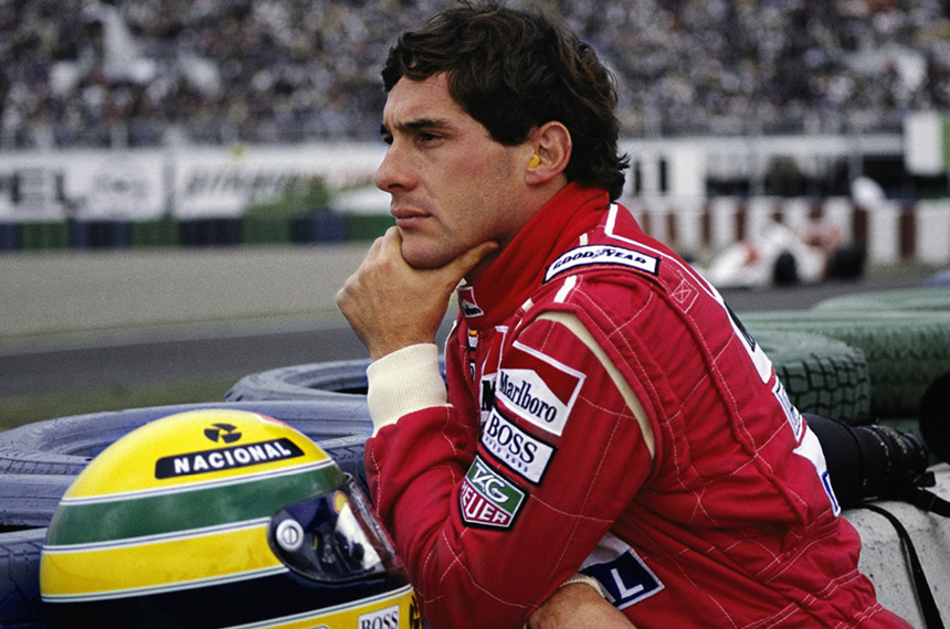 Ayrton Senna vestindo macacão de corrida vermelho, com a mão no queixo, observando a pista e, na sua frente, seu capacete amarelo.