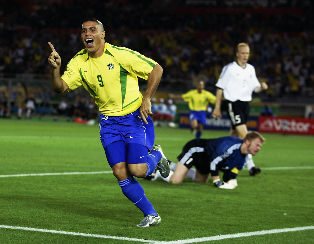 Icônica imagem de Ronaldo Fenômeno na Copa de 2002, correndo em campo e apontando para cima, comemorando gol marcado. Ao fundo, Oliver Khan, goleiro da Alemanha, fita o fundo do gol. Ronaldo veste camisa amarela e short azul, uniforme da Seleção Brasileira.