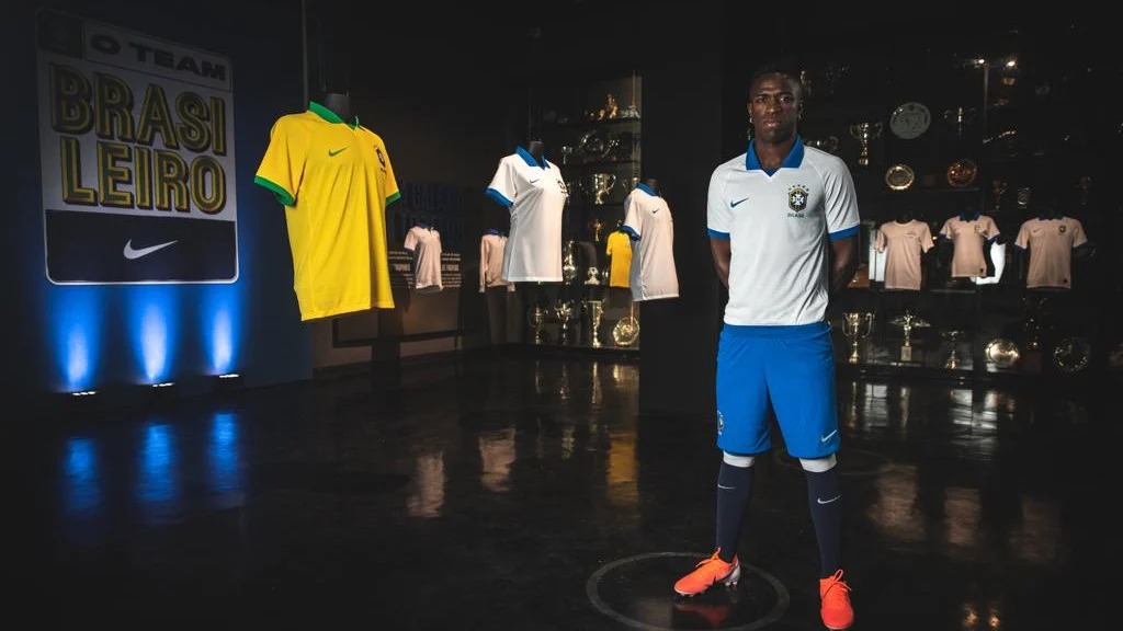 Vinícius Jr usa uniforme branco e azul do Brasil, posando em museu, perto de outros manequins com camisas do Brasil.