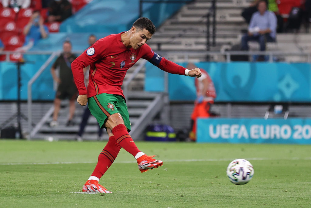 Cristiano Ronaldo em campo pela Seleção de Portugal, vestindo camisa vermelha e short verde, enquanto chuta a bola, usando chuteiras laranjas.