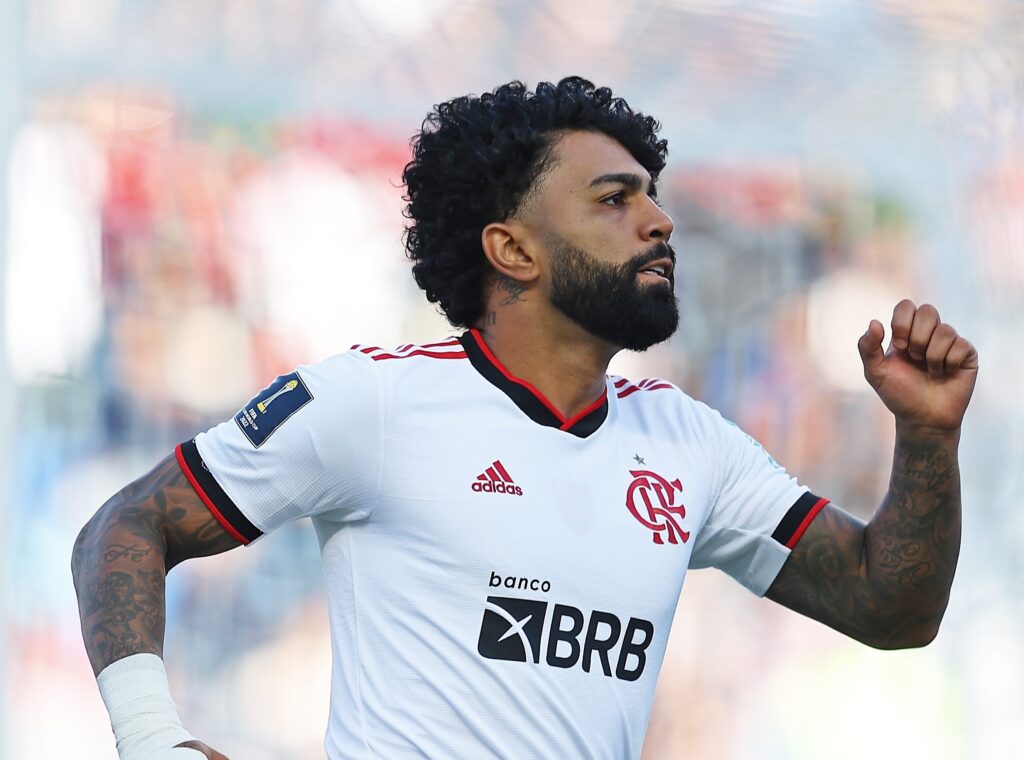 Gabigol viaja com Flamengo. Jogador aparece com camisa branca com detalhes em preto e vermelho. Ele está correndo com as mãos para o alto.