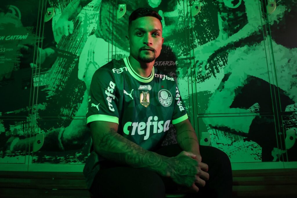 Palmeiras contrata Artur, de volta ao clube após 3 anos no Bragantino - Artur posa para foto, sentado, com um mural do Palmeiras atrás dele, junto com uma iluminação verde na foto