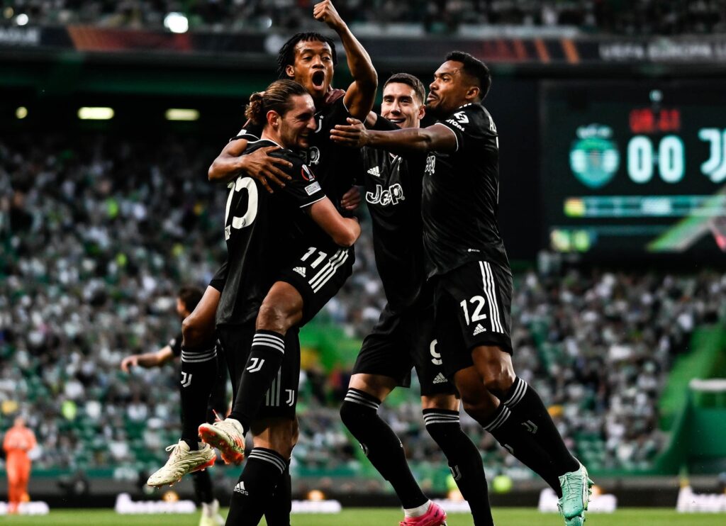 Juventus vai à semifinal da Liga Europa. Quatro jogadores se abraçam felizes ao comemorarem um gol. Eles vestem uniforme preto com detalhes em branco.