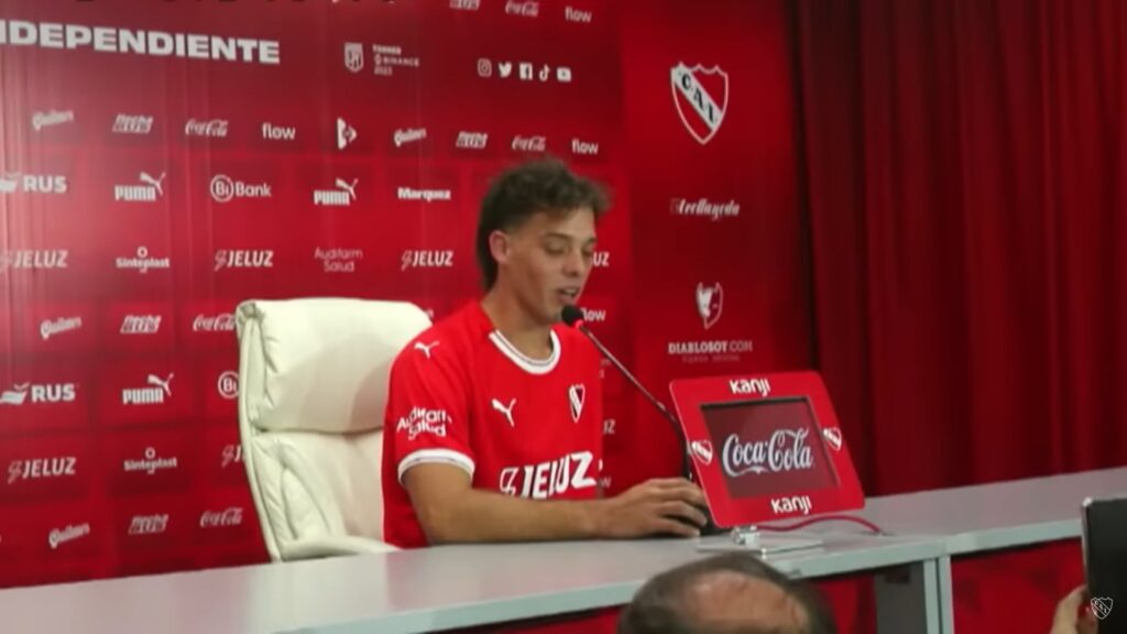 Independiente vive crise. Homem está sentado em uma cadeira branca em frente a um microfone. Ele está usando uma camiseta vermelha em detalhes brancos.