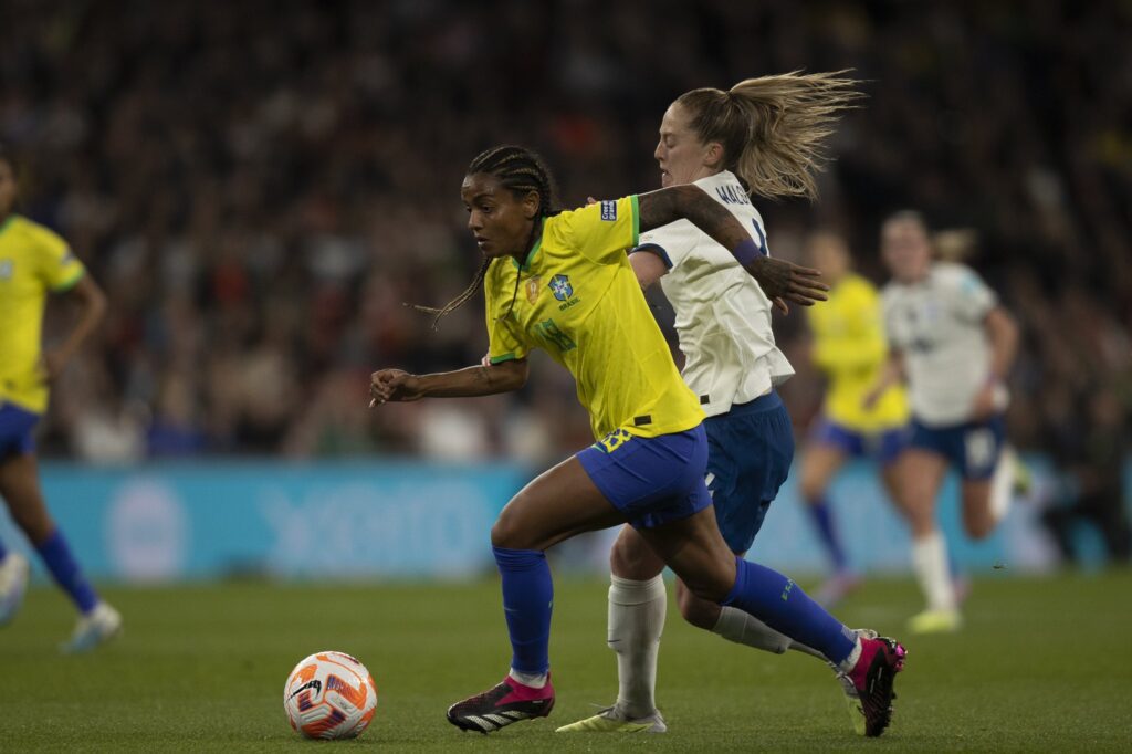 Seleção Brasileira perde. Duas jogadoras aparecem correndo atrás da bola. Elas estão no primeiro plano da imagem, uma veste uniforme amarelo e azul. A outra veste uniforme branco.