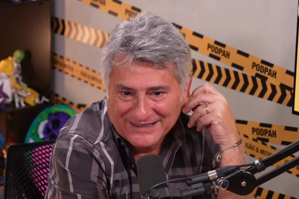 Cléber Machado saiu da Globo: Narrador revela que quase pediu demissão antes da Copa do Mundo - Cléber Machado, sentado de frente para o microfone no estúdio do PodPah, falando com um sorriso tímido no rosto, enquanto segura a orelha