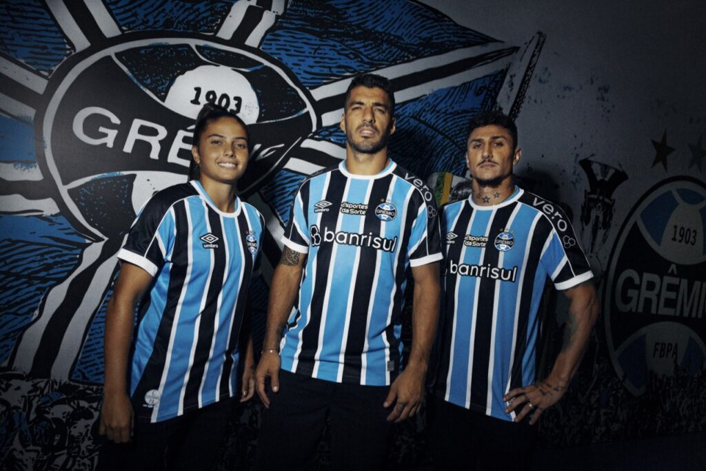 Nova camisa do Grêmio para 2023 é lançada; confira as fotos - jogadores do Grêmio posam para foto de divulgação da nova camisa do Grêmio, ao fundo uma parede com o escudo do Grêmio pintado.