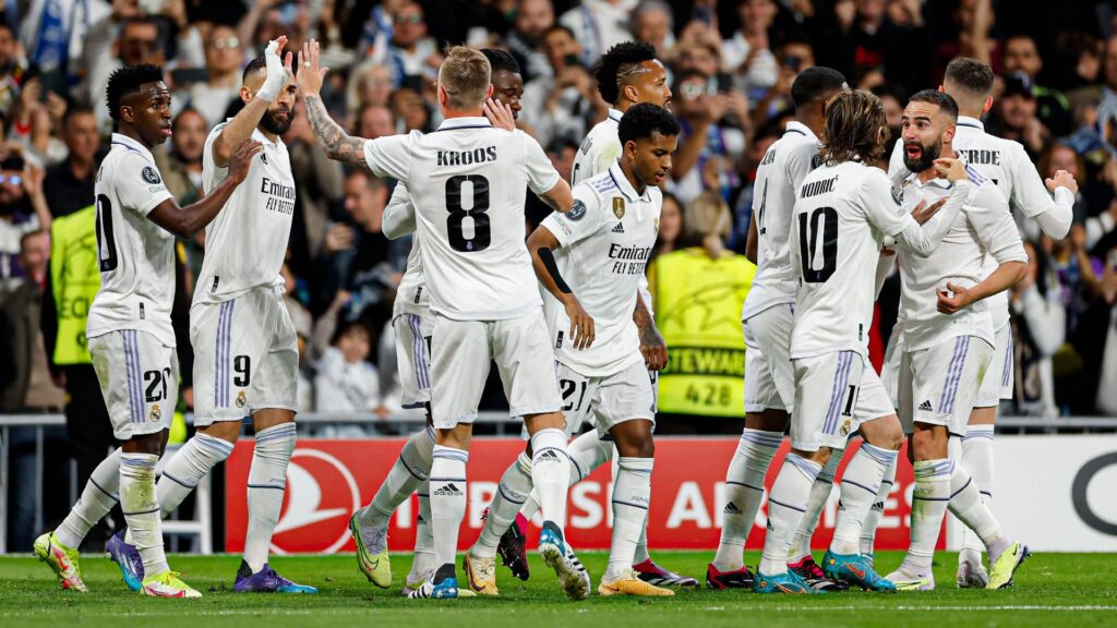 Real Madrid vence Chelsea. Jogadores aparecem se cumprimentando após o gol. A maioria aparece de costas, e vestem uniforme branco com detalhes em preto.
