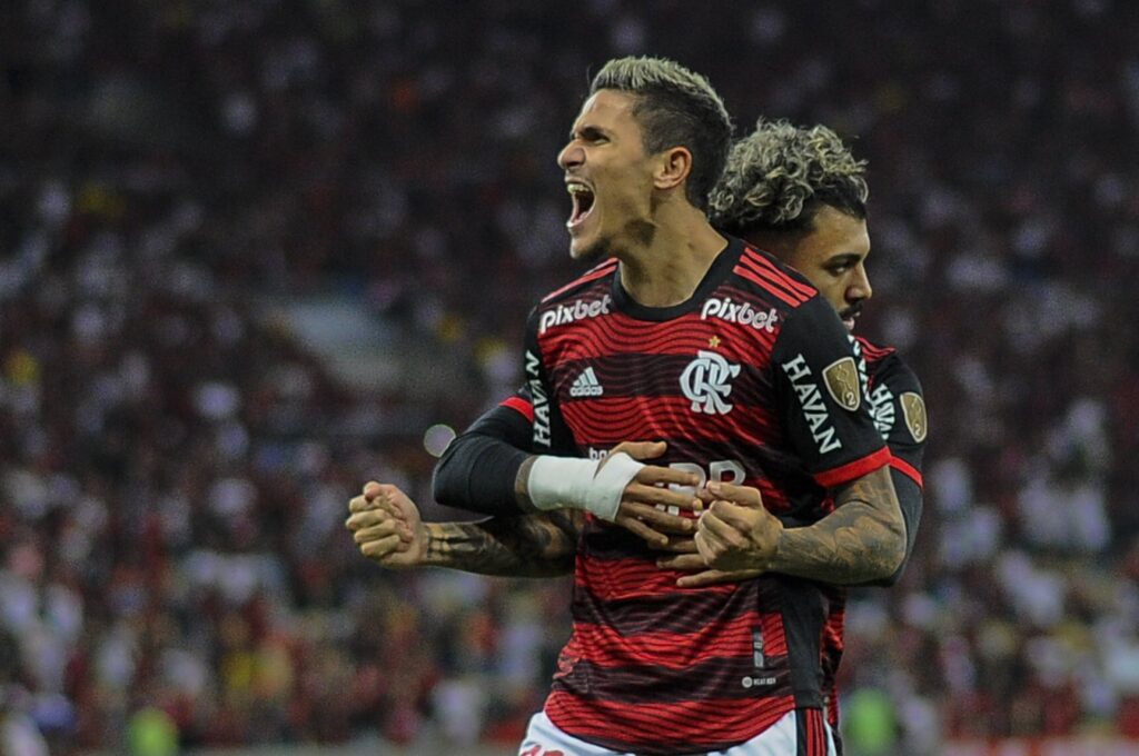 Pedro desfalca o Flamengo: Dois jogadores aparecem se abraçando. Pedro está em mais evidencia e aparece com os braços levantados e gritando. Gabigol está o abraçando de costas.