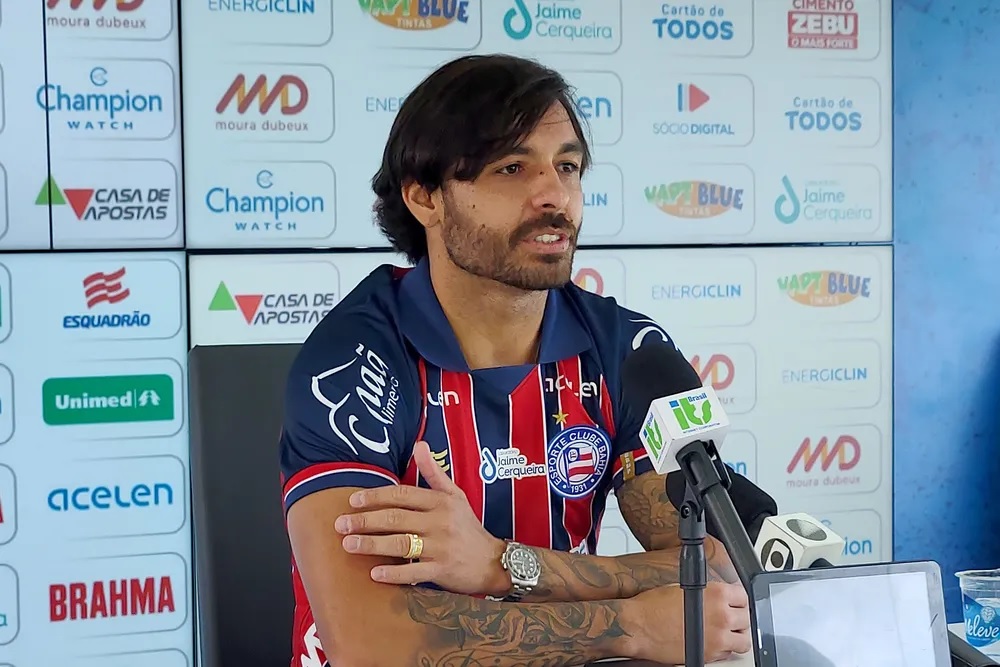 Jogador está sentado diante a um microfone. Ele está dando entrevista coletiva e veste camisa do Bahia, azul com detalhes em vermelho e branco.