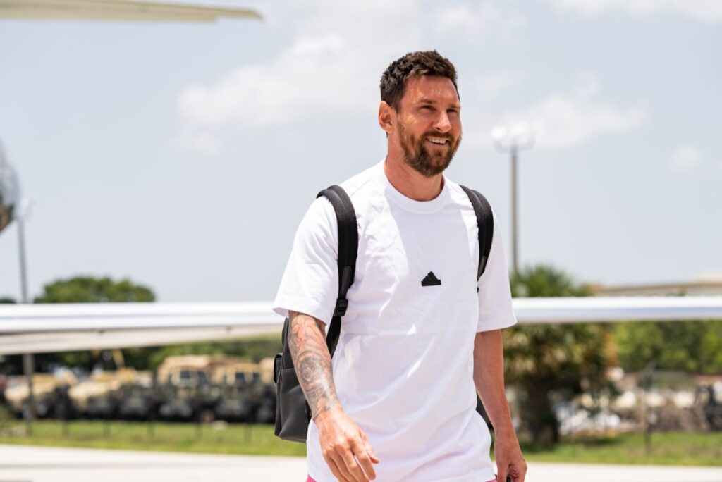 Messi vai ser emprestado pelo Inter Miami? Jornalista revela planos do craque - Messi usa uma camisa branca com a logo da adidas ao centro dela, enquanto usa uma mochila nas costas e caminha sorridente