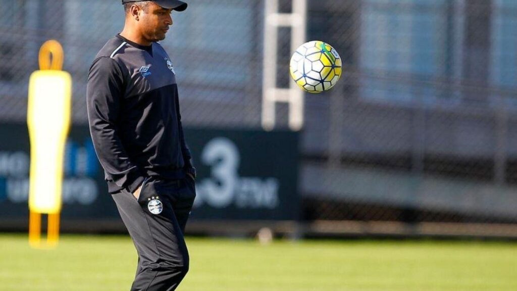 Roger Machado, enquanto era treinador do Grêmio, se prepara para dominar uma bola que vem em sua direção. Ele está com agasalho preto de treino do Grêmio, em um campo no centro de treinamento do clube gaúcho