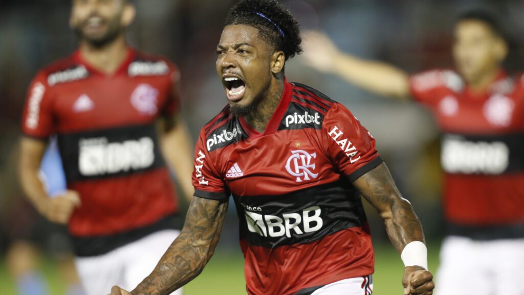 Gabigol viaja com o Flamengo. Jogador aparece em primeiro plano, com uma camisa vermelha com detalhes em preto e branco. Ele está gritando ao comemorar um gol.