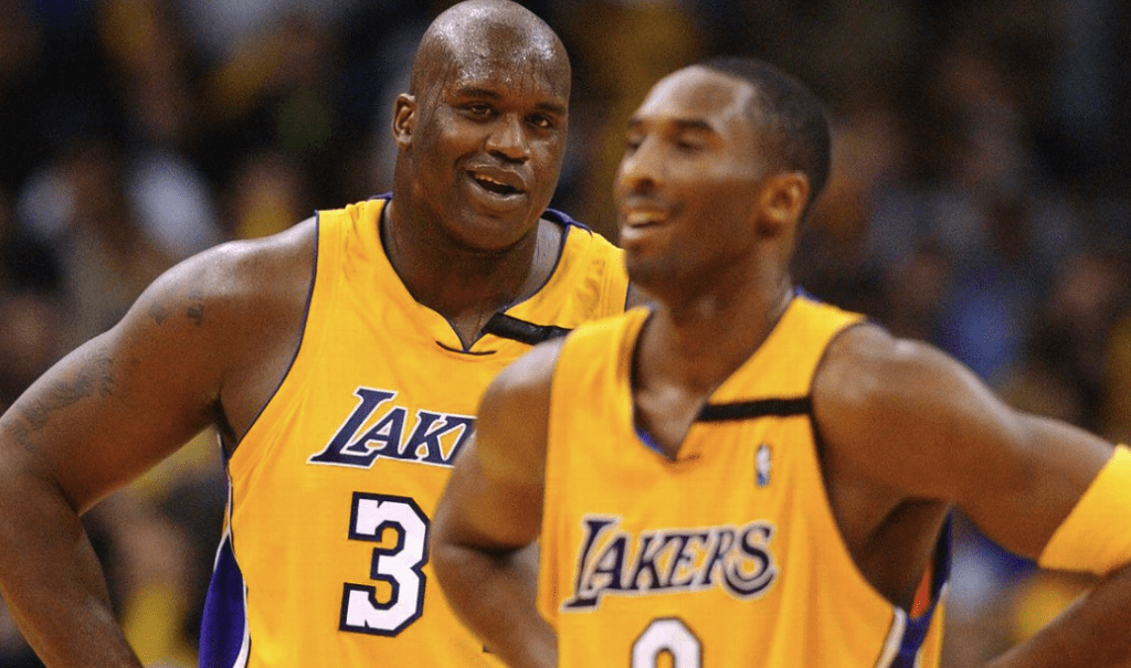 Shaquille O'Neal em quadra defendendo a camisa dos Lakers. O Jogador está sorrindo e bastante suado pelo jogo