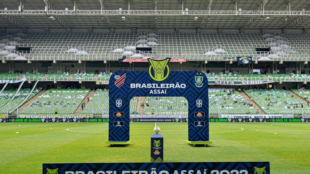 Placa com a frase "Brasileirão Assaí 2022" em frente a totem com a bola do campeonato e, ao fundo, arco da competição, indicando o duelo entre Athletico-PR e América-MG na Arena da Baixada.