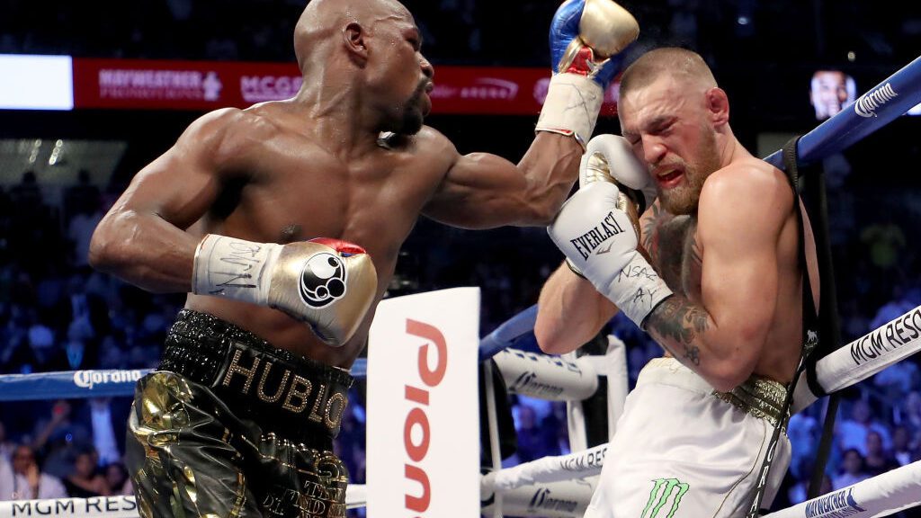 Dentro de ringue de boxe, Floyd Mayweather golpeando Conor McGregor. Ambos usam luvas e shorts de boxe.