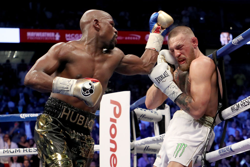 Dentro de ringue de boxe, Floyd Mayweather golpeando Conor McGregor. Ambos usam luvas e shorts de boxe.