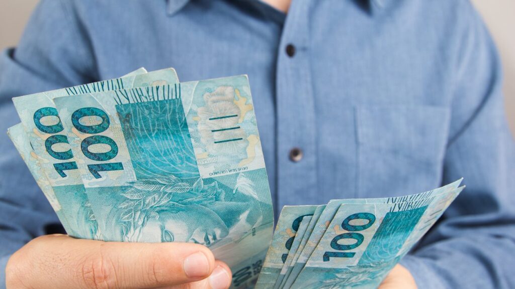 Homem vestindo camisa azul, com botões, contando notas de 100 reais