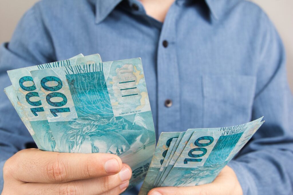 Homem vestindo camisa azul, com botões, contando notas de 100 reais