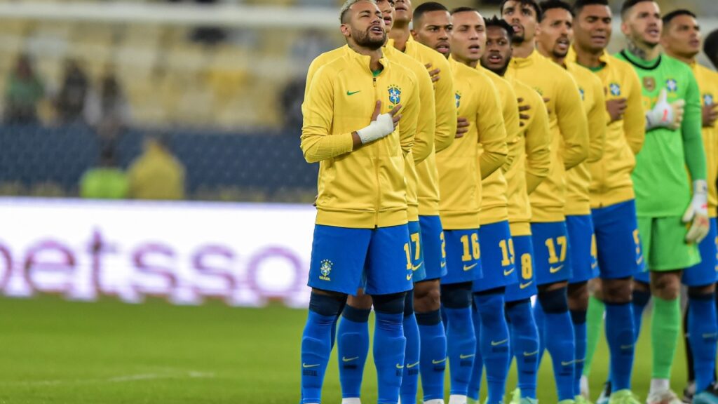 Seleção Brasileira perfilada em campo, jogadores com a mão no peito, entoando o hino nacional
