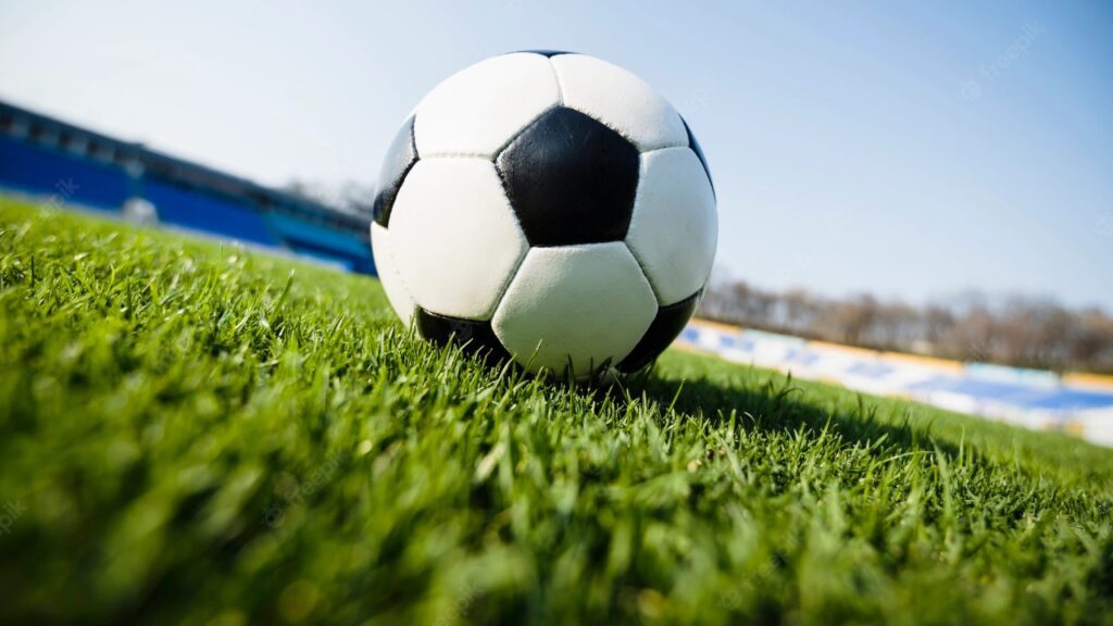 Bola de futebol preta e branca, em cima de gramado de campo de futebol, em imagem levemente inclinada.