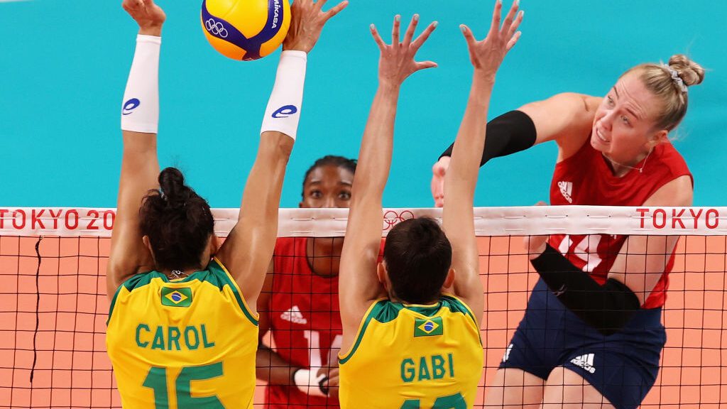 Jogadora de vôlei corta a bola por cima da rede e adversárias subindo para bloquear