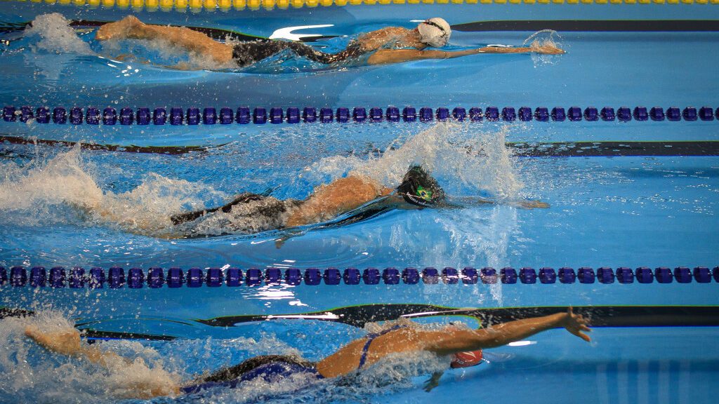 Três nadadoras durante prova em piscina, cada uma em uma raia