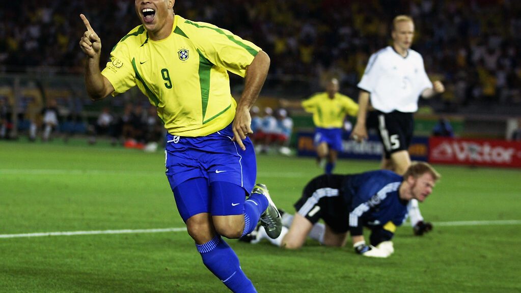 Icônica imagem de Ronaldo Fenômeno na Copa de 2002, correndo em campo e apontando para cima, comemorando gol marcado. Ao fundo, Oliver Khan, goleiro da Alemanha, fita o fundo do gol. Ronaldo veste camisa amarela e short azul, uniforme da Seleção Brasileira.