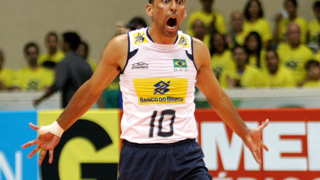 Serginho Escadinha em quadra pela Seleção Brasileira de vôlei. De braços abertos, andando pra a frente e com expressão de comemoração raivosa, Serginho usa short azul escuro e camisa branca.