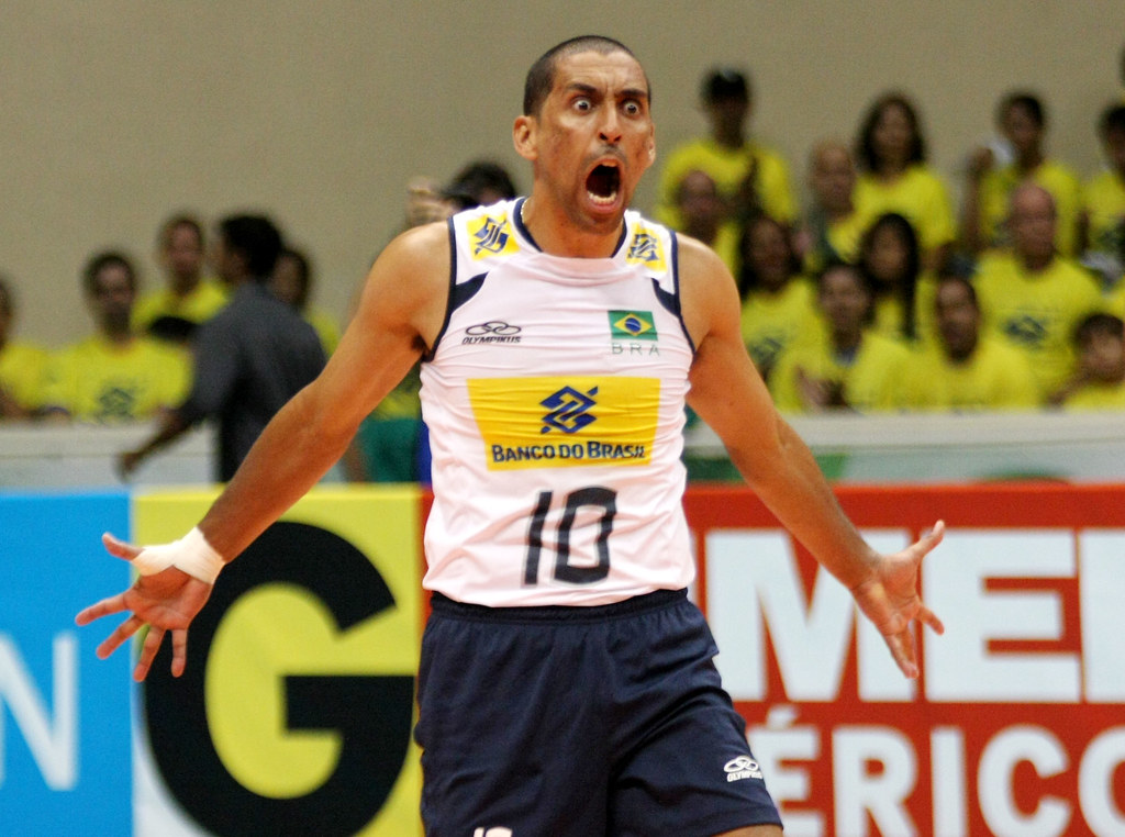 Serginho Escadinha em quadra pela Seleção Brasileira de vôlei. De braços abertos, andando pra a frente e com expressão de comemoração raivosa, Serginho usa short azul escuro e camisa branca.