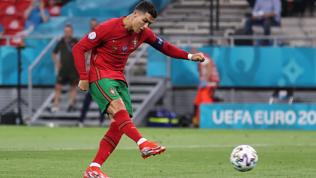 Cristiano Ronaldo em campo pela Seleção de Portugal, vestindo camisa vermelha e short verde, enquanto chuta a bola, usando chuteiras laranjas.