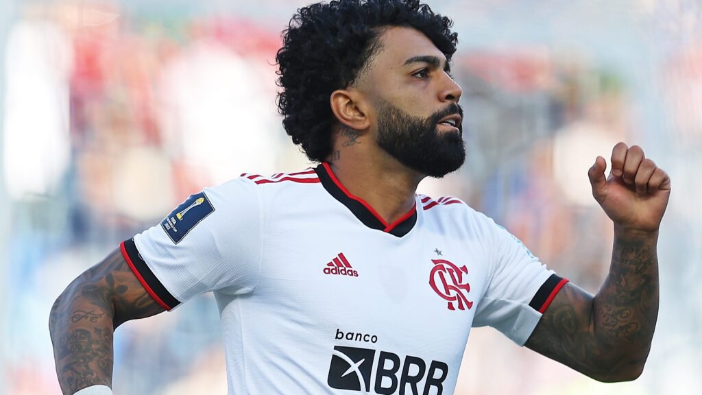 Gabigol viaja com Flamengo. Jogador aparece com camisa branca com detalhes em preto e vermelho. Ele está correndo com as mãos para o alto.
