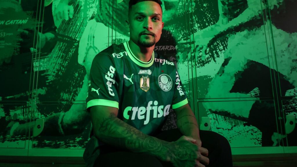 Palmeiras contrata Artur, de volta ao clube após 3 anos no Bragantino - Artur posa para foto, sentado, com um mural do Palmeiras atrás dele, junto com uma iluminação verde na foto
