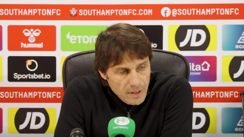 Antonio Conte aparece de camisa preta e concedendo entrevista coletiva. Ele está concentrado falando em frente a um microfone verde.