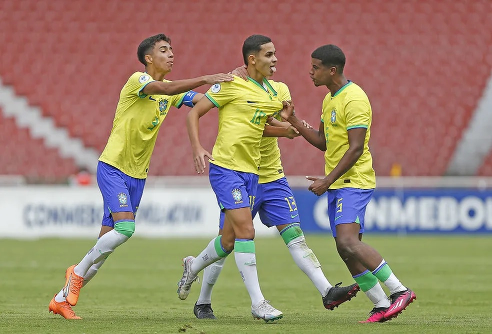 Seleção Brasileira vence Paraguai. Três garotos aparecem na imagem. Eles estão comemorando gols e vestem uniforme com camiseta amarela em detalhes em verde, e shorts azul com detalhes em branco.