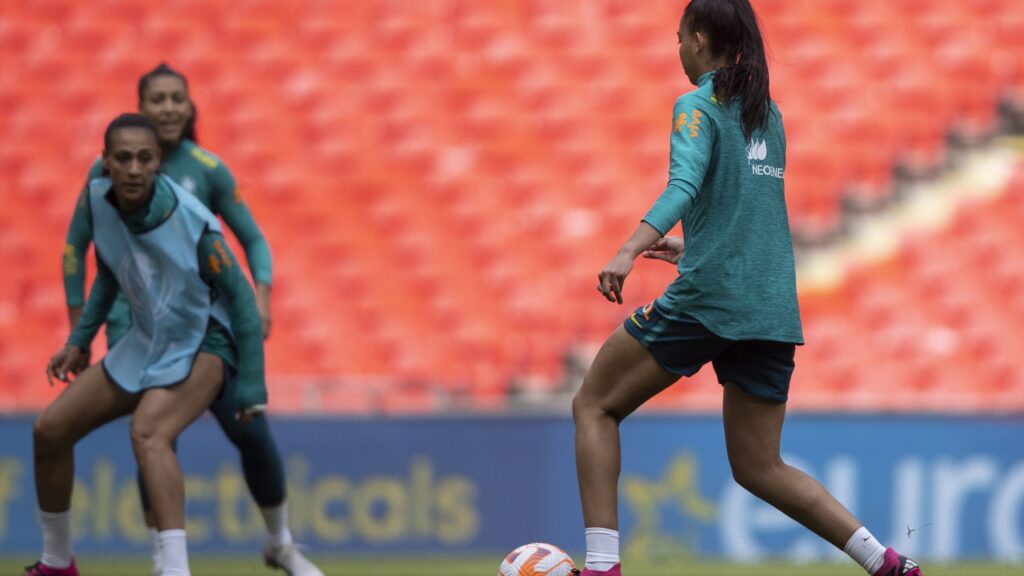Seleção Brasileira faz último treino. Duas jogadoras aparecem em campo com a bola nos pés. Elas estão de costas e vestem uniformes azuis.
