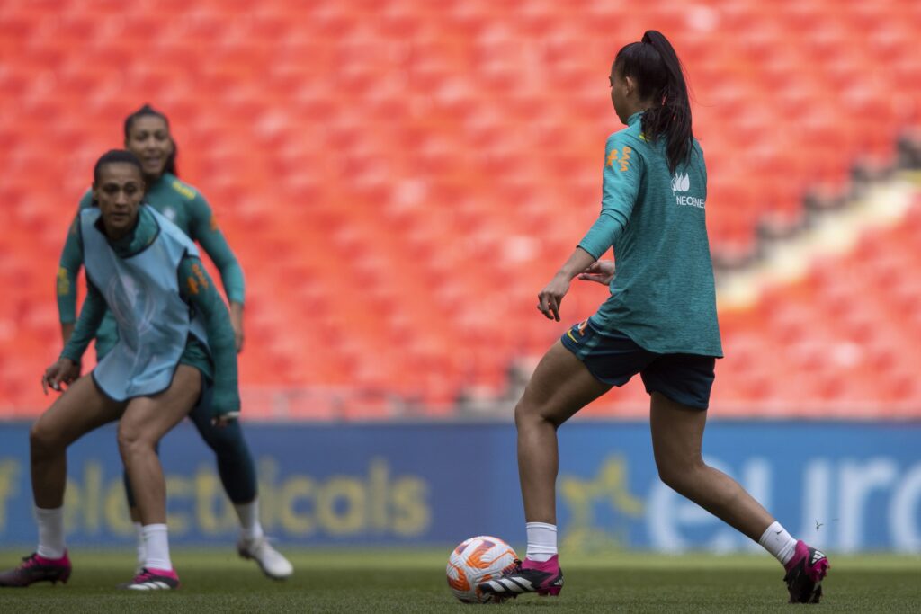 Seleção Brasileira faz último treino. Duas jogadoras aparecem em campo com a bola nos pés. Elas estão de costas e vestem uniformes azuis.