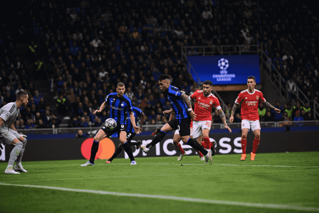 Inter de Milão empata com Benfica. Na imagem, quatro jogadores aparecem no segundo plano da imagem. Eles estão correndo atrás da bola. Os dois mais próximos da bola vestem uniforme azul com listras verticais pretas. Os outros dois estão um pouco atrás e vestem uniforme vermelho com detalhes em branco.