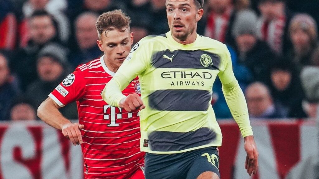 Manchester City empata com Bayern. Dois jogadores aparecem em foco. No primeiro plano, um corre na frente e veste uniforme amarelo com listras grossas pretas. O outro aparece logo atrás, também correndo, e veste uniforme vermelho em detalhes em brancos.