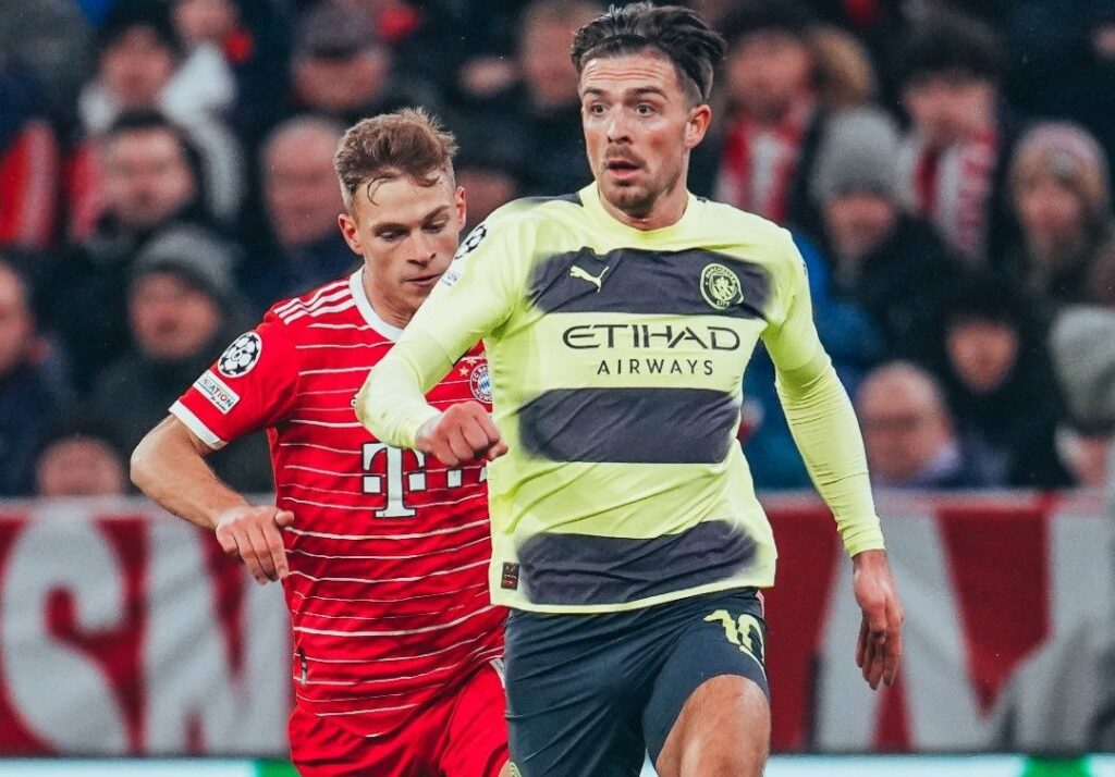 Manchester City empata com Bayern. Dois jogadores aparecem em foco. No primeiro plano, um corre na frente e veste uniforme amarelo com listras grossas pretas. O outro aparece logo atrás, também correndo, e veste uniforme vermelho em detalhes em brancos.