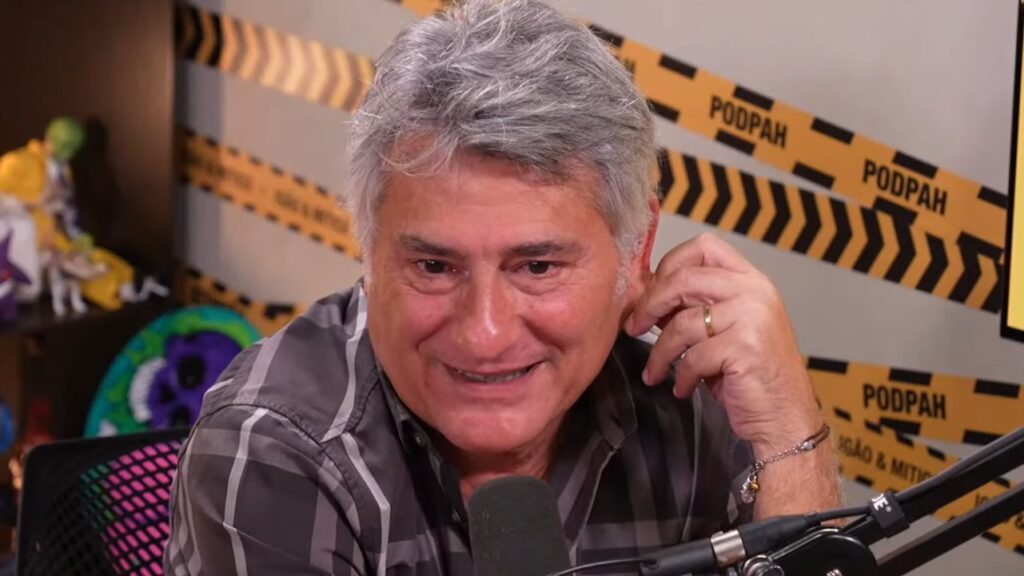 Cléber Machado saiu da Globo: Narrador revela que quase pediu demissão antes da Copa do Mundo - Cléber Machado, sentado de frente para o microfone no estúdio do PodPah, falando com um sorriso tímido no rosto, enquanto segura a orelha